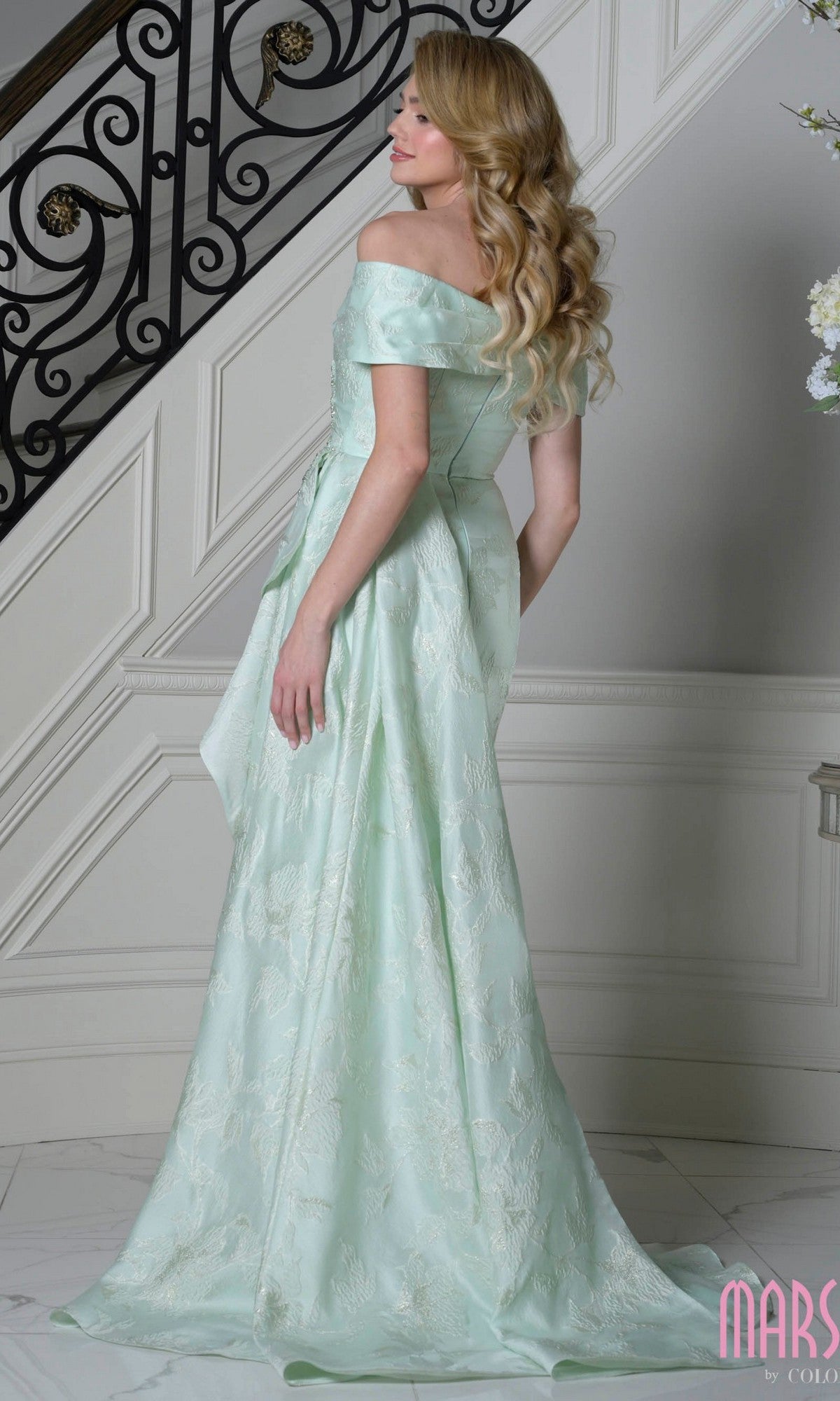 A woman wearing the Marsoni MV1401 long formal dress in light green stands on a marble floor near an ornate staircase, facing away from the camera.